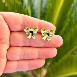 May include: A pair of bow-shaped stud earrings. The earrings feature a green and yellow design with black outlines. The earrings are held in a person's hand against a blurred green background.