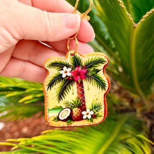 May include: A tropical-themed keychain featuring a palm tree, flowers, a pineapple, and a coconut. The keychain has a gold-colored clasp and ring. The background is a yellow gradient with a brown border.