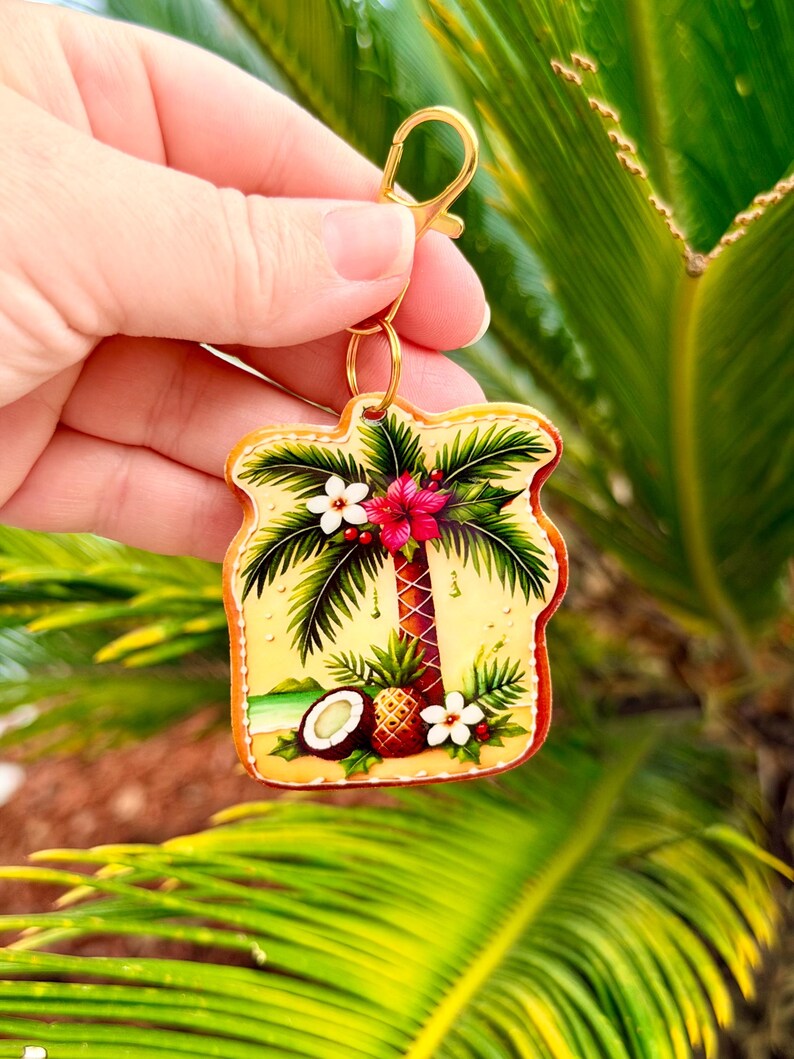 May include: A tropical-themed keychain featuring a palm tree, flowers, a pineapple, and a coconut. The keychain has a gold-colored clasp and ring. The background is a yellow gradient with a brown border.