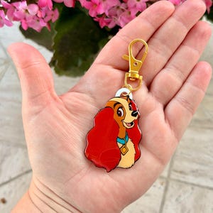 May include: A Lady and the Tramp keychain featuring Lady. The keychain has a gold-colored clasp and a charm with a cartoon image of Lady, with red hair, a blue collar, and a gold tag. The background includes pink flowers.