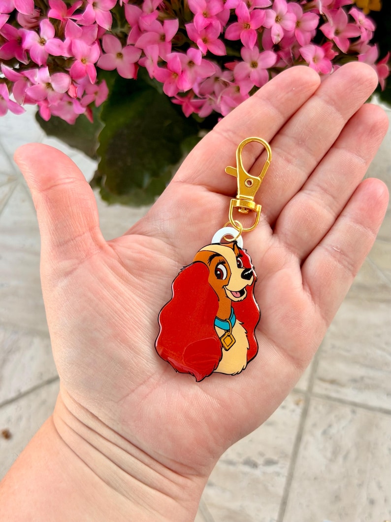 May include: A Lady and the Tramp keychain featuring Lady. The keychain has a gold-colored clasp and a charm with a cartoon image of Lady, with red hair, a blue collar, and a gold tag. The background includes pink flowers.