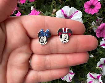 Può includere: Un paio di piccoli orecchini a bottone in legno con facce di personaggi dei cartoni animati. Un orecchino raffigura un personaggio con un fiocco blu e l'altro con un cappello rosso, bianco e blu. Gli orecchini sono tenuti in una mano su uno sfondo di fiori rosa e bianchi.