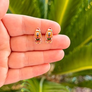 May include: A pair of small stud earrings featuring a cartoon dog character with light brown fur, large ears, and a black nose. The dog is wearing a red and white Santa hat and a green bow tie, rendered in a flat, colorful enamel style design.