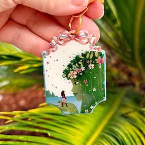 K&ouml;nnte beinhalten: Ein rechteckiger Schl&uuml;sselanh&auml;nger mit einem goldenen Verschluss zeigt eine farbenfrohe Illustration. Die Illustration zeigt eine Frau mit gr&uuml;nen Haaren und einem Blumenkranz und eine andere Frau am Strand. Der Schl&uuml;sselanh&auml;nger ist mit einer rosa Schleife und kleinen blauen Blumen verziert.