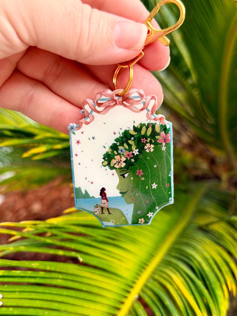 K&ouml;nnte beinhalten: Ein rechteckiger Schl&uuml;sselanh&auml;nger mit einem goldenen Verschluss zeigt eine farbenfrohe Illustration. Die Illustration zeigt eine Frau mit gr&uuml;nen Haaren und einem Blumenkranz und eine andere Frau am Strand. Der Schl&uuml;sselanh&auml;nger ist mit einer rosa Schleife und kleinen blauen Blumen verziert.