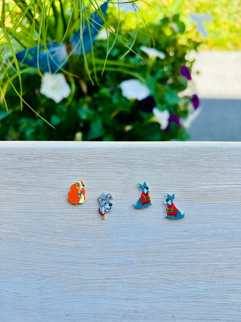 May include: Four colorful dog-themed embellishments. One is a light brown dog with orange hair, a second is a gray dog with a red collar, and two are blue dogs wearing red and green sweaters. They are arranged on a light gray surface.