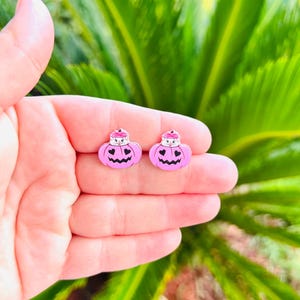 May include: A pair of pink pumpkin-shaped earrings with black carved-out faces and small white hats. The earrings are held in a hand against a backdrop of green foliage, perfect for Halloween.