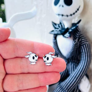 May include: A pair of white Zero stud earrings with black eyes and an orange nose. The earrings are held in a hand, with a Jack Skellington figure in the background. The earrings are a fun accessory for fans of the movie.