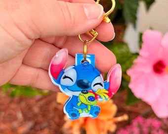May include: A blue and white Stitch keychain with a gold-colored clasp. The keychain features a cartoon character holding a small doll. The background includes colorful flowers.