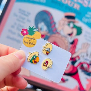 May include: A set of four cartoon character enamel pins on a white card. The pins feature a parrot wearing a sombrero, a duck, and a rooster, all with yellow and red accents. The card has a pineapple-shaped topper with text that reads "Pineapples & Pixie Dust co."