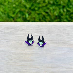 Puede incluir: Un par de pendientes con una imagen de dibujos animados de Mal&eacute;fica de La Bella Durmiente de Disney. Los pendientes son negros, morados y verdes.