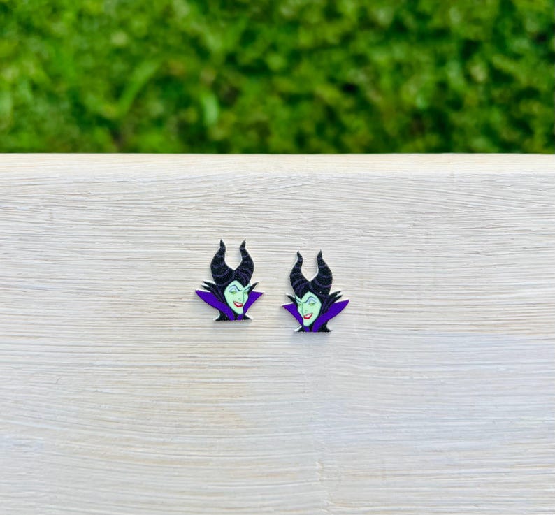 Puede incluir: Un par de pendientes con una imagen de dibujos animados de Mal&eacute;fica de La Bella Durmiente de Disney. Los pendientes son negros, morados y verdes.