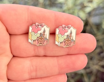 May include: Two rectangular enamel pins featuring a pink castle, Mickey Mouse ears, and a pink bow. The pins have a gold border and are held in a hand against a blurred background.
