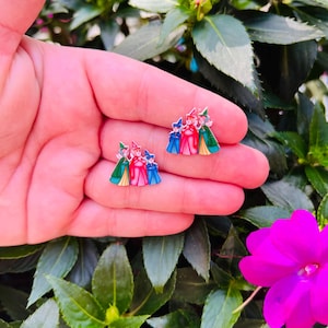 May include: A pair of colorful enamel earrings featuring a design of three figures in red, blue, and green outfits. The earrings are set against a background of green foliage and a pink flower.