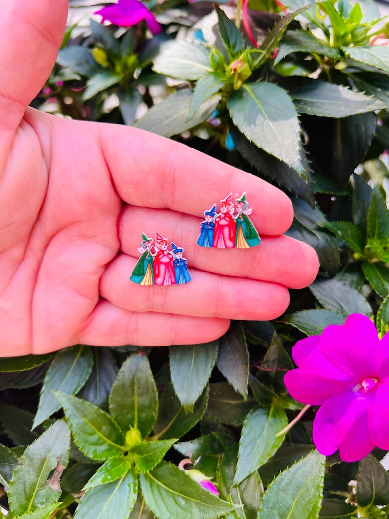 May include: A pair of colorful enamel earrings featuring a design of three figures in red, blue, and green outfits. The earrings are set against a background of green foliage and a pink flower.