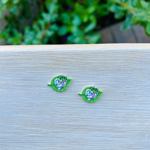 May include: Two small, whimsical earrings. Each earring features a cartoon-style insect with a crown, set on a green leaf shape. The insects have blue, purple, and white details. The background is a light wood surface.