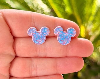May include: A pair of Mickey Mouse-shaped stud earrings. The earrings feature a blue and pink firework-like pattern. The earrings are held in a hand against a blurred green background.