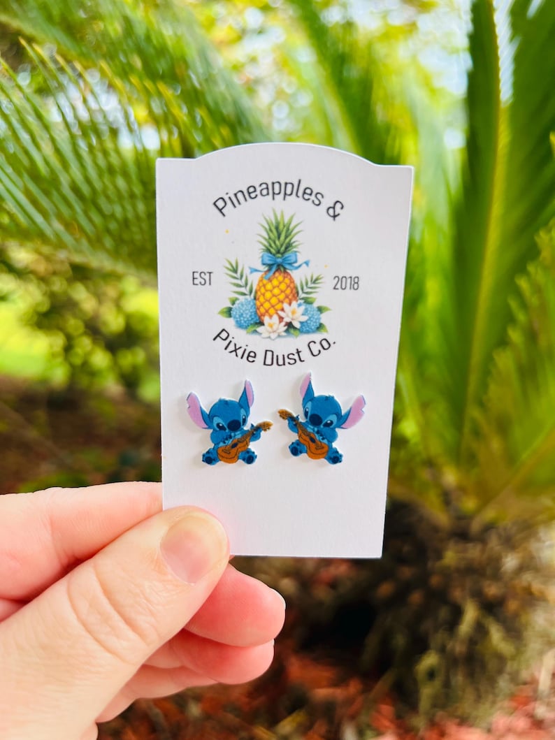 May include: Two blue Stitch earrings, each playing a ukulele, are presented on a white card. The card displays the text "Pineapples & Pixie Dust Co." alongside a pineapple illustration. The background shows blurred green leaves.