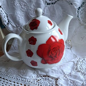 Può includere: Teiera in ceramica bianca decorata con grandi e piccoli motivi a rosa rossa. La teiera ha un manico curvo, un beccuccio e un coperchio. Poggia su una tovaglietta di pizzo bianco.