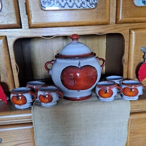 Può includere: Un servizio da tè in ceramica a tema mela, composto da una teiera con coperchio e sei tazzine. Il set presenta una combinazione di colori bianco e rosso con un design rustico. Il set è esposto su un panno di iuta.