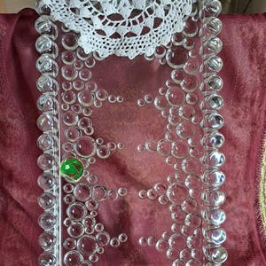 Könnte beinhalten: Ein rechteckiges Tablett aus klarem Glas mit einem Blasendesign an den Rändern und auf der Oberfläche. Oben auf dem Tablett befindet sich eine weiße Spitzendecke. In der Mitte des Tabletts befindet sich ein grünes und goldenes Dekorationselement.