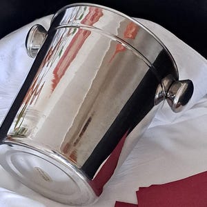 Può includere: Un secchiello per il vino in acciaio inossidabile lucido con due manici arrotondati. Il secchiello è cilindrico con una parte superiore leggermente svasata. È appoggiato su un panno bianco con un tovagliolo bordeaux. Il secchiello riflette l'ambiente.