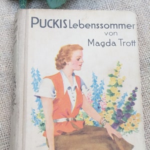 Könnte beinhalten: Ein Vintage-Buch mit dem Titel "Puckis Lebenssommer von Magda Trott" mit einem weißen Einband und einer bunten Illustration einer Frau, die in einem Blumenfeld sitzt. Das Buch ist im "Titania-Verlag Stuttgart" erschienen.