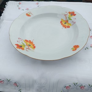 Peut inclure: Bol en céramique blanc avec un bord festonné et un bord doré. Le bol est orné de motifs floraux orange et jaune. Le bol est posé sur une nappe blanche.