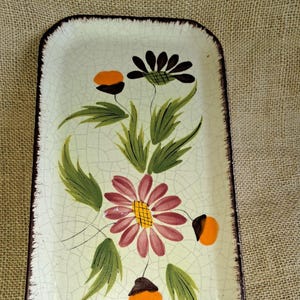 Puede incluir: Bandeja rectangular de cerámica con diseño floral. La bandeja tiene un fondo color crema con acabado craquelado y borde marrón oscuro. Las flores son en tonos rosa, azul, naranja y negro, con hojas verdes.