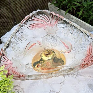 Könnte beinhalten: Eine Servierschale aus klarem Glas mit einem dekorativen rosa-weißen Blumendesign am Rand. Die Schale hat einen goldfarbenen Metallsockel und steht auf einer weißen Spitzendecke. Die Schale ist wahrscheinlich Vintage.
