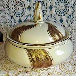 Op de afbeelding: Crèmekleurige keramische serveerschaal met deksel, met goudkleurige accenten. De schaal heeft een afgeronde vorm met een deksel met een decoratieve handgreep. De gouden accenten zijn in een golvend patroon. Een vintage stuk.