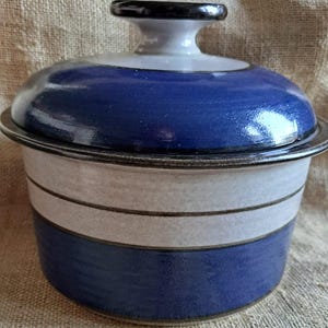 Op de afbeelding: Een blauwe en witte keramische voorraadpot met een deksel. De pot heeft een zwarte rand en een witte streep rond het midden.