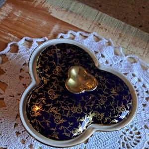 Könnte beinhalten: Eine blaue und goldene Keramik-Schmuckschatulle in Form eines Kleeblatts mit einem goldenen Herzen in der Mitte. Die Schatulle steht auf einer weißen Häkeldeckchen.