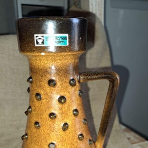 Peut inclure: Un vase en céramique brun avec une anse et une surface texturée. Le vase porte une étiquette qui indique "Gärtnerei Helm".