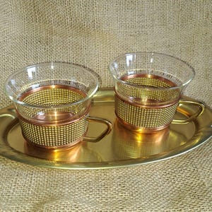 Puede incluir: Dos tazas de té de vidrio transparente con fundas y asas de malla metálica de color cobre, apoyadas sobre una bandeja de metal dorado. La bandeja tiene forma ovalada y curvada. El fondo es una tela texturizada de color beige.