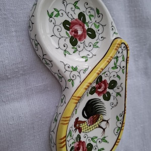 Peut inclure: Un repose-cuillère en céramique blanche en forme de goutte, orné d'un motif peint à la main représentant un coq, des roses rouges et des vignes vertes. Les bords présentent une bordure jaune avec des traits bruns et une finition brillante.