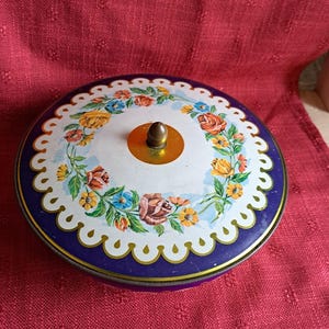 Può includere: Scatola rotonda vintage con un design floreale. Il coperchio presenta uno sfondo bianco con una ghirlanda di fiori colorati, un bordo smerlato e un pomello in ottone. La scatola ha un bordo blu scuro e si trova su una superficie rossa.