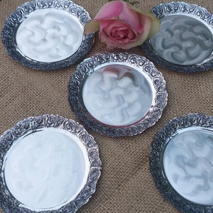 Può includere: Cinque sottobicchieri argentati con un design floreale ornato. I sottobicchieri sono rotondi e hanno un bordo leggermente rialzato. Sono disposti in cerchio su una superficie di juta.