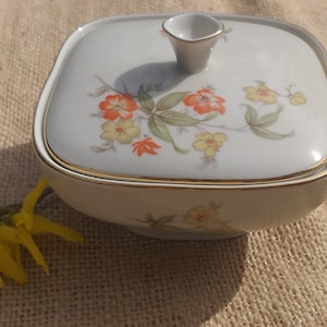 Könnte beinhalten: Eine weiße Porzellan-Zuckerdose mit Deckel, verziert mit einem Blumendesign mit orangefarbenen, gelben und grünen Blumen. Die Schale hat einen goldenen Rand.