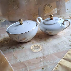 Op de afbeelding: Een witte porseleinen suiker- en roomstel met gouden accenten. De suikerpot heeft een deksel met een gouden knop en beide stukken zijn versierd met een bloemmotief. De set staat op een goud-beige patroon oppervlak.