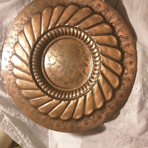 Könnte beinhalten: Ein großes, rundes Serviertablett aus Kupfer mit einem komplizierten, geprägten Design. Das Tablett hat eine erhöhte Mitte mit einem kleineren, kreisförmigen Design.