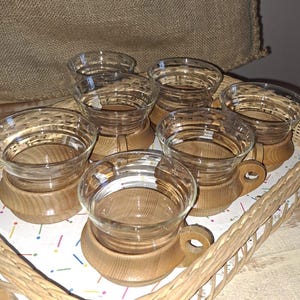 Könnte beinhalten: Satz von sechs klaren Glasbechern mit Holzgriffen und -böden, angeordnet in einem Weidenkorb auf einer Holzoberfläche.