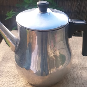 Może przedstawiać: Srebrny czajnik z czarnym uchwytem i pokrywką. Czajnik stoi na powierzchni z juty.