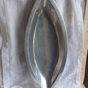 Könnte beinhalten: Eine silberfarbene, blattförmige Servierschale. Die Schale hat eine glatte, reflektierende Oberfläche und ein spitzes Ende. Sie liegt auf einem weißen, strukturierten Tuch. Die Schale wird wahrscheinlich zum Servieren von Speisen oder als Dekorationsartikel verwendet.