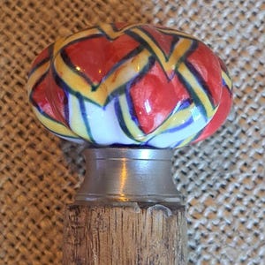 Pode incluir: Uma bengala de madeira com uma cabeça de cerâmica. A cabeça é decorada com um padrão geométrico em vermelho, amarelo e azul.