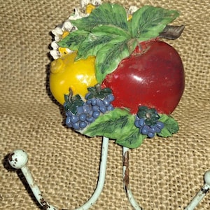 Könnte beinhalten: Ein dekorativer Wandhaken mit einem roten Apfel, einer gelben Zitrone und blauen Trauben mit grünen Blättern. Das Obst ist an einem weißen Metallhaken mit zwei gebogenen Armen befestigt. Der Hintergrund ist ein strukturierter, beigefarbener Stoff.