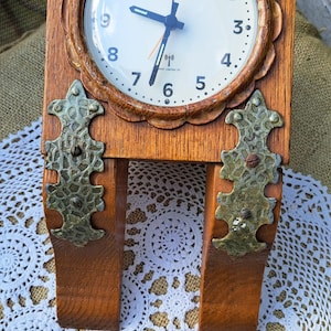 May include: A wooden clock with a white face and black hands. The clock has a decorative design around the edge and the words "IDEEN WELT" printed on the face. The clock is mounted on a wooden stand with two decorative metal pieces.
