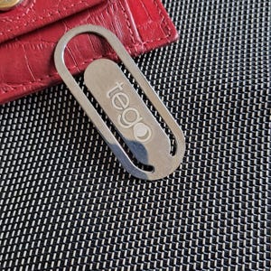 May include: A red, textured leather wallet with a gold button and a silver metal bookmark. The bookmark has the word "tego" printed on it. The background is a black and white patterned surface.