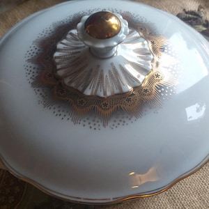 Może przedstawiać: Biała porcelanowa miska do serwowania z pozłacanym brzegiem i misternym wzorem w kolorze złotym. Pokrywka ma złoty uchwyt.