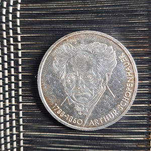 Può includere: Una moneta d'argento raffigurante Arthur Schopenhauer, con gli anni 1788-1860 incisi. La moneta è rotonda e presenta un aspetto leggermente usurato. È visibile anche la scritta "ARTHUR SCHOPENHAUER". La moneta è su una superficie nera strutturata.
